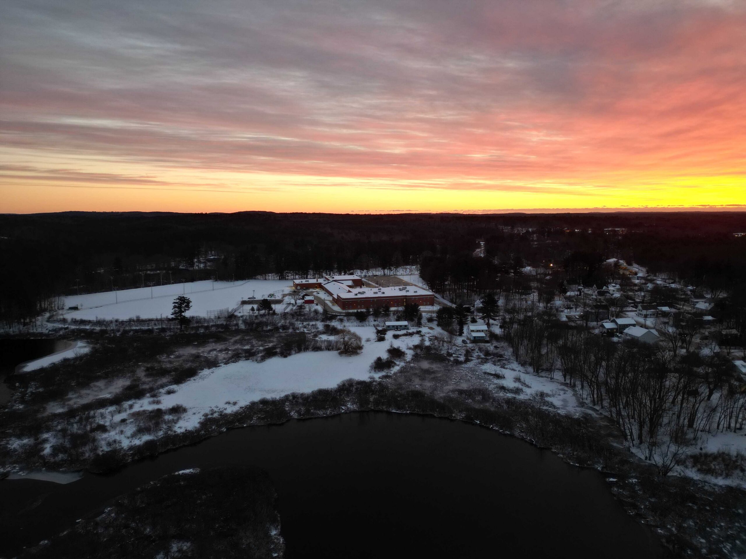 Winter sunset aerial over Raymond, New Hampshire, with a river and snowy neighborhood below